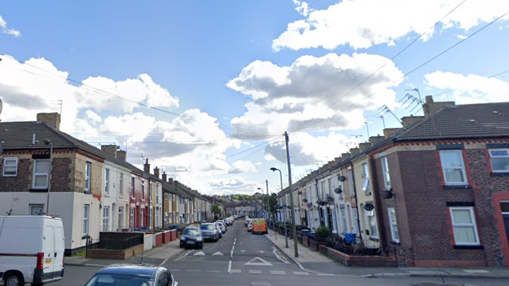 A van and a car at a road junction with a street of terraced houses behind