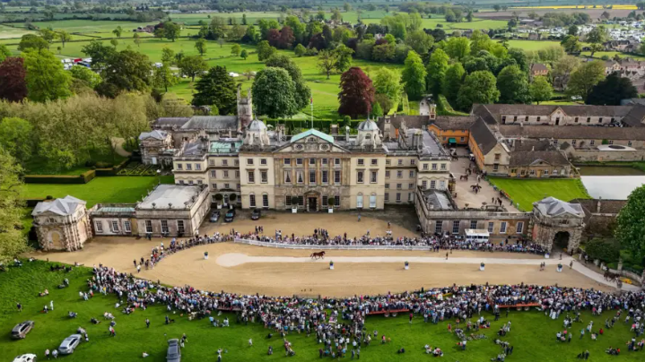 Stately home Badminton House with crowds in front of it and trees and fields behind it. It is a drone view up and looking down at the front of the house.