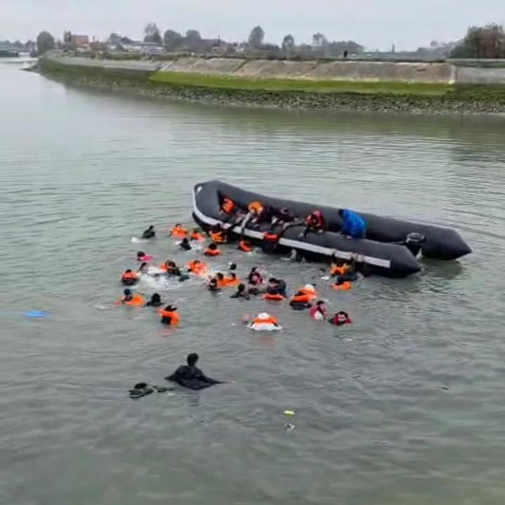 People, mostly wearing orange life jackets, swim to a black dinghy in a canal.