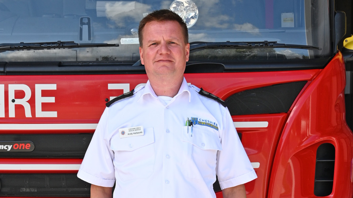 A man in a white shirt with the words Cheshire Fire and Rescue emblazoned over one pocket stands in front of a red fire engine.