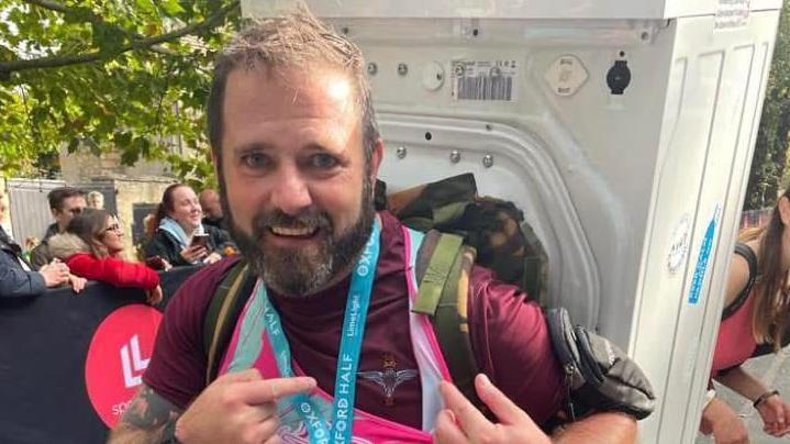 Martin has short brown hair and a brown beard. He is at the end of his half marathon, with a washing machine strapped to his back. He is pointing to a medal around his neck.