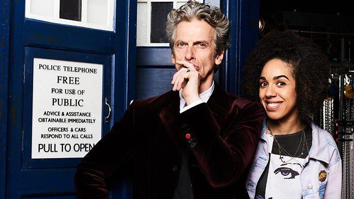 Peter Capaldi, with wavy grey hair wearing a burgundy velvet blazer and white shirt and holding his hand over his face in a puzzled manner with a frowning expression on his face, is on the left of the picture. Beside him is a woman in a black and white t-shirt and denim overshirt. She has black afro hair and is smiling at the camera. Behind them is a blue phone box, known as the Tardis.