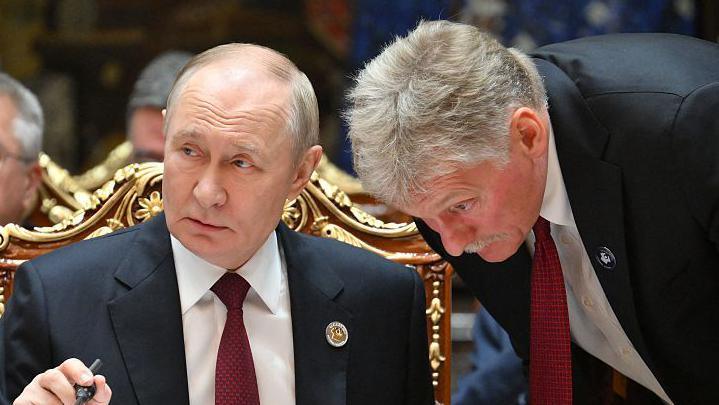 Russian President Vladimir Putin in a suit and red tie say behind a table, Peskov, a man with grey hair wearing a suit is bending down listening to him talk.