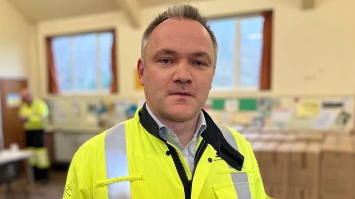 A man with short, dark hair looking at the camera. He is wearing a yellow high-vis coat.