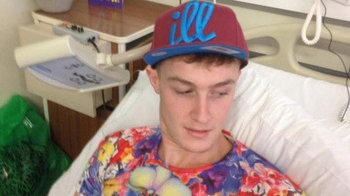 Ryan Finan in a brightly coloured patterned T-shirt is laying in a hospital bed. He has a baseball cat with the word "ill" written on in cursive blue font.