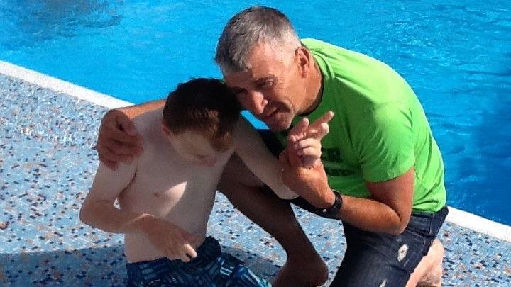 Ryan wearing blue swimming trunks sitting on the edge of a pool and looking down. Beside him, Richard is kneeling down with one arm around Ryan and the other holding his wrist. Richard is wearing blue denim shorts and a bright green T-shirt. 