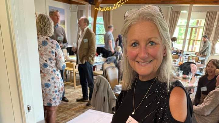 A woman with long grey hair is smiling to the camera. She has reading glasses on her head and is wearing a dark blue dress. She is standing in a hotel conference room with people behind her. 