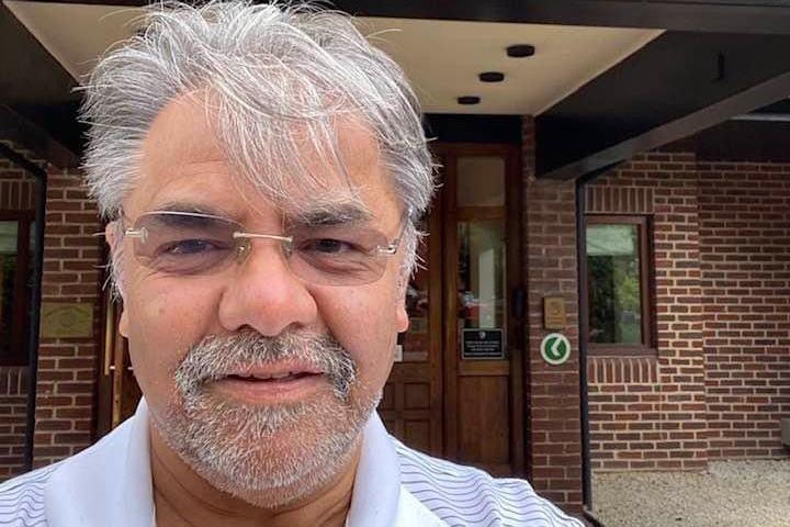 A selfie of a man with white hair and a white goatee beard looking at the camera. He wears frameless glasses. A red bricj building is behind him. He wars a blue and white stripy polo shirt.