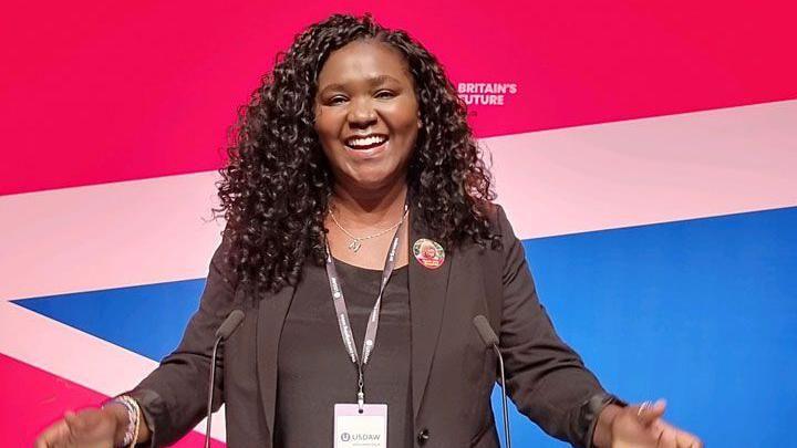 Marianne Kimani with curling hair falling to her shoulders standing at a lectern 