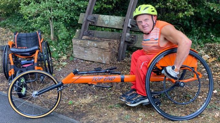 A picture of a man in an orange racing wheelchair, wearing orange and a yellow helmet. He is pictured at the side of a path in a park with a second smaller orange wheelchair pictured in the background.