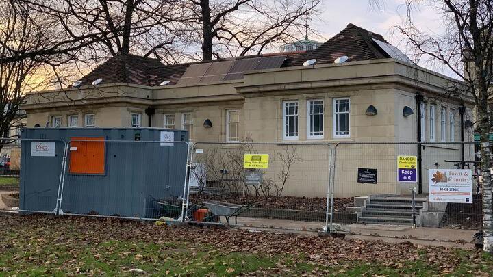 The public toilet building, made from pale yellow stone, with metal fencing around it, and a small grey container for workers. Grass and leaves are in the foreground.