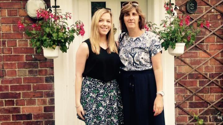 A woman with blonde hair stands outside a front door with an older woman with brown hair on a summer's day