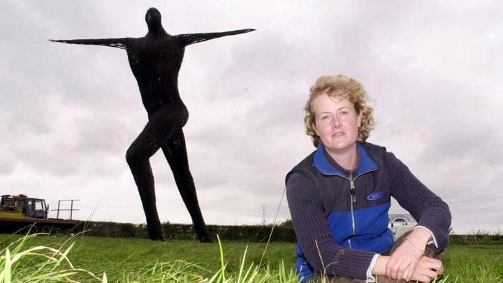 A wide shot of he fully intact Willow Man, with his arms outstretched in a field, and in the foreground is artist Serena De La Hay, sat on the floor looking at the camera.