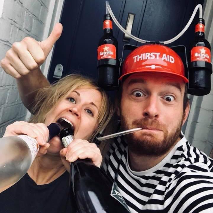 Tom Price wears a red “Thirst Aid” beer hat fitted with bottles and straws, while Beth pretends to drink directly from two wine bottles. They pose together outside their front door.
