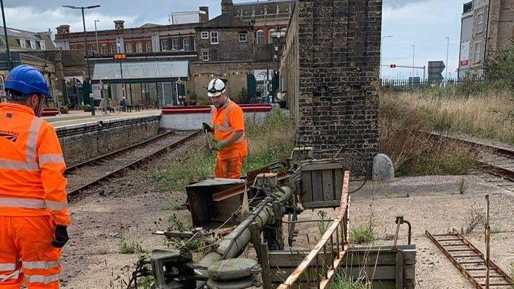 Two Network Rail workers wearing orange high visibility suits and helmets are dismantling old signal equipment close to railway lines at the station