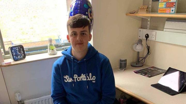 Logan Wareham sat in his student accommodation. He is wearing a blue hoody that says San Portabalo in white bubble writing. He is sat at his desk with a blue balloon behind him. 