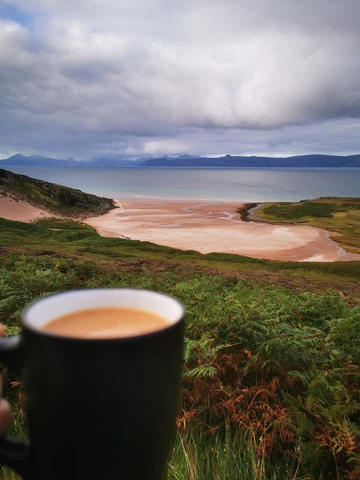 A cup of tea in the foreground with a sprawl of grass and beach in front looking out to the sea under a cloudy sky