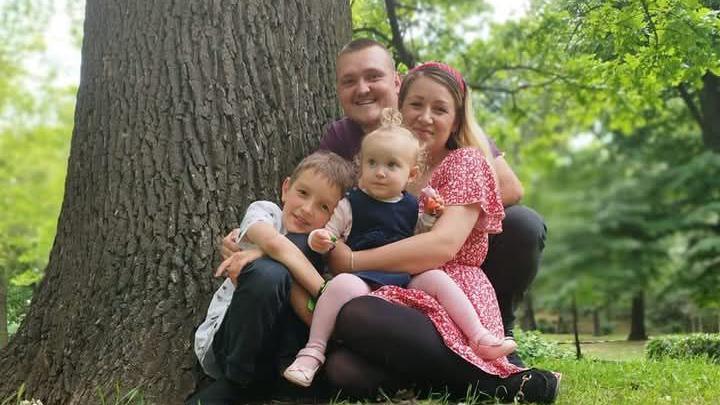 Family photo of Lewis and Jordan Vye with their son and young daughter. They are sitting at the base of a tree hugging each other and smiling at the camera.