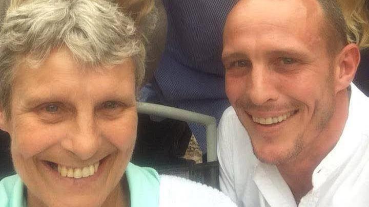 A middle aged woman with greying, short hair, pictured with a young man who looks like her son. They are both smiling.