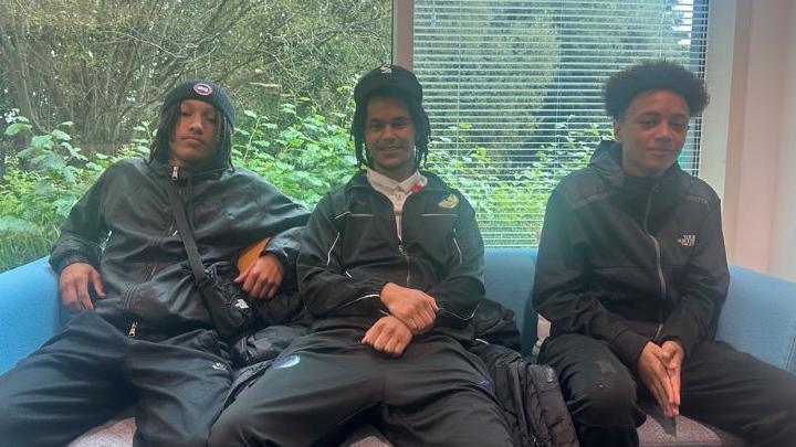 Three teenage boys - Roman Shance, Kartel Forde and Shaelon - sitting on a blue sofa each wearing black trousers and jackets. Two of the boys are wearing hats