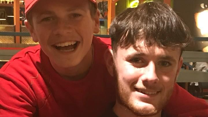 A boy in a red jumper and red cap smiles and hugs another older boy with brown messy hair and a beard. 