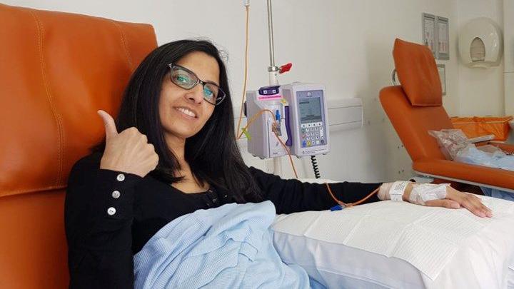 A woman in a black top sitting in an orange hospital chair with a blue blanket and receiving hospital treatment. She is smiling and giving a thumbs-up.