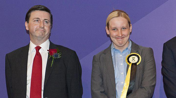 Douglas Alexander with a sad face is wearing a dark suit, white shirt and red tie and has a red rose as a buttonhole. He is standing next to Mhair Black who has a happy face. She is wearing a grey jacket and blue shirt. she is wearing a yellow and black SNP rosette