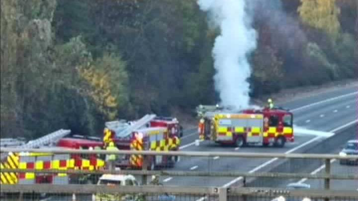 A traffic camera image above the M11 carriageway. Three fire engines block the road. Smoke from a car on fire can be seen billowing upwards from behind one of the fire engines.