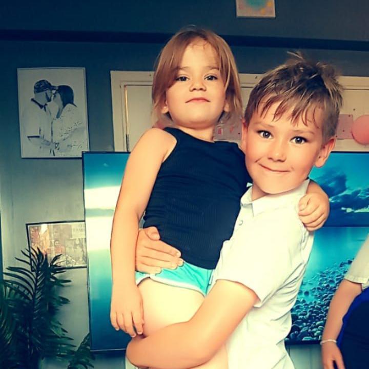 Boy with short brown hair wearing a white t-shirt carrying a girl with shoulder length brown hair wearing a dark top and blue shorts. There are pictures behind them on a blue wall. There is a TV and a plant.