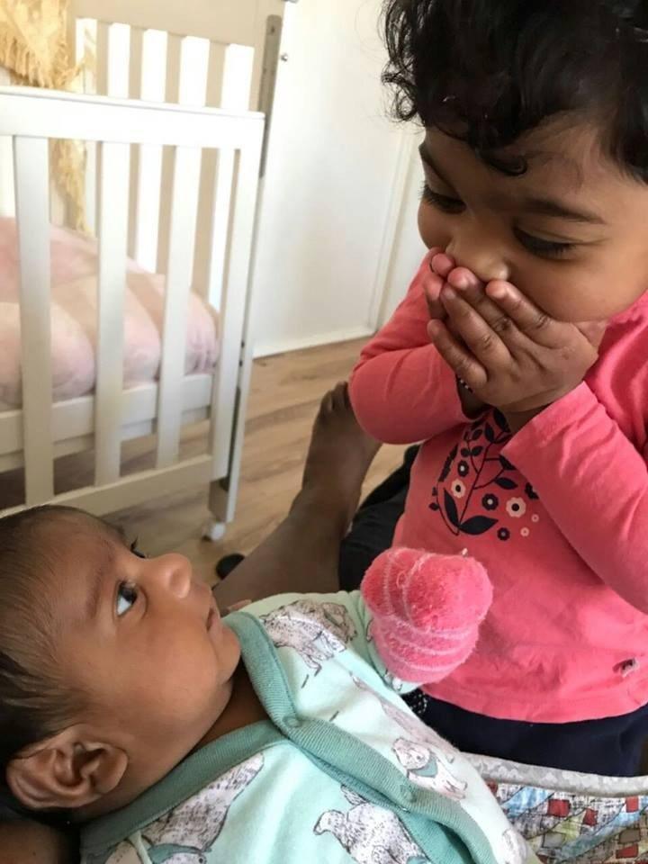 Two-year-old Kopiga, with her hands covering her mouth, looks down at her sister nine-month-old Dharuniga