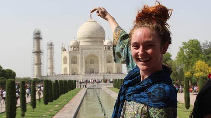 A young woman is posing in front of the Taj Mahal. She's standing down the lawn that leads up to the entrance with her finger pointed at the top of the building. She has wavy auburn hair tied up in bun and is wearing a green top with a blue and black scarf over her shoulders. Small thin trees are lined up along a fountain leading up to the building. The ivory coloured building has rotundas on the roof.