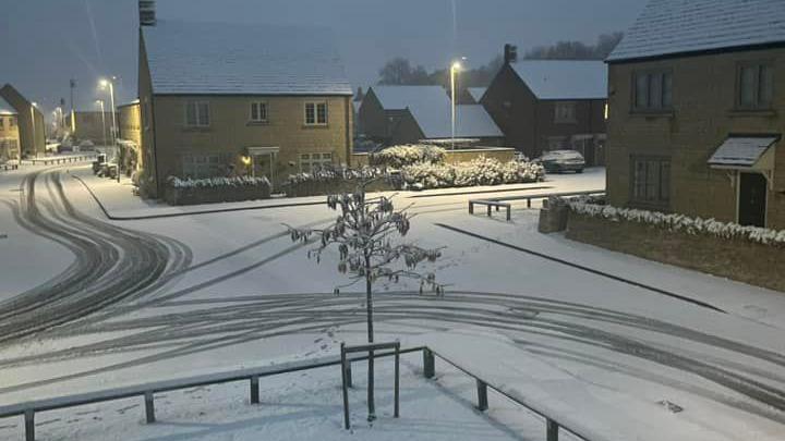 A view out of a second storey bedroom window onto the street below. It is a wide road with houses on each corner. The roofs are covered in snow and the streetlights are on, lighting up the dim scene. There are swirling patterns left in the snow from car tyres.