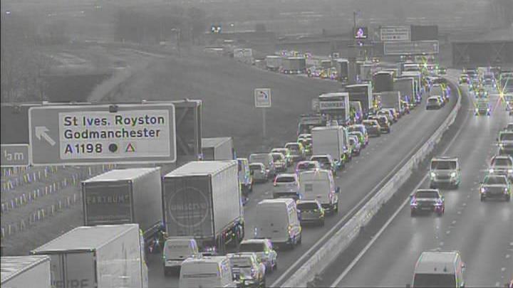 A black and white CCTV image showing the A14. The left lane is blocked by traffic. There is a large sign to the left of the road pointing left for St Ives, Royston and Godmanchester. 