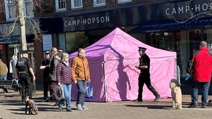 Pink tent covers the bench where Paddington Bear statue was on Northbrook Street. Officers are seen talking to a few people. Other residents are seen walking dogs and looking at the tent. It's a sunny day.