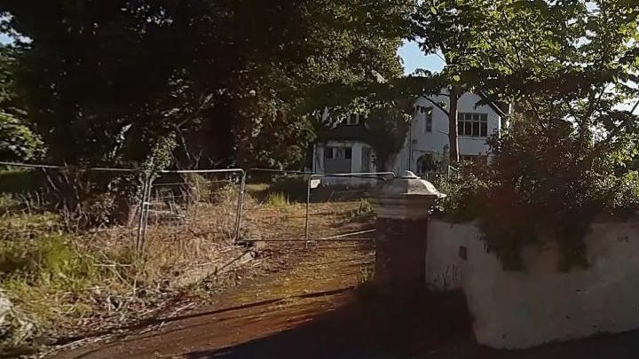 St Olave's House, which is a large white house with a black roof and traditional house windows. It has an overgrown garden with large trees at the entrance, which has safety fencing across it to keep people off the site.