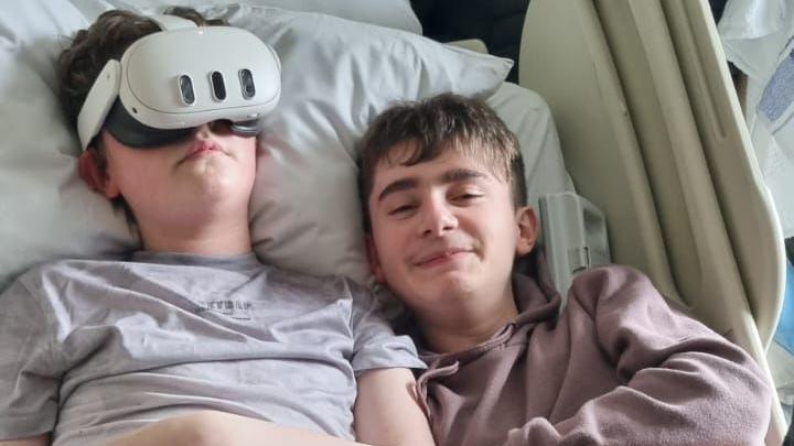 Kyle wearing a grey t-shirt and with his VR headset on in a hospital bed. Lying next to him is his brother Liam, who is smiling at the camera wearing a light brown hoodie. 