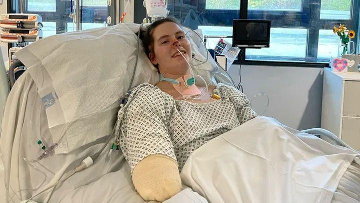 Lily is lying in a hospital bed, wearing a hospital gown. She has tubes up her nose and on her chest. Her right arm has been amputated to the elbow and has been bandaged. She is smiling at the camera.