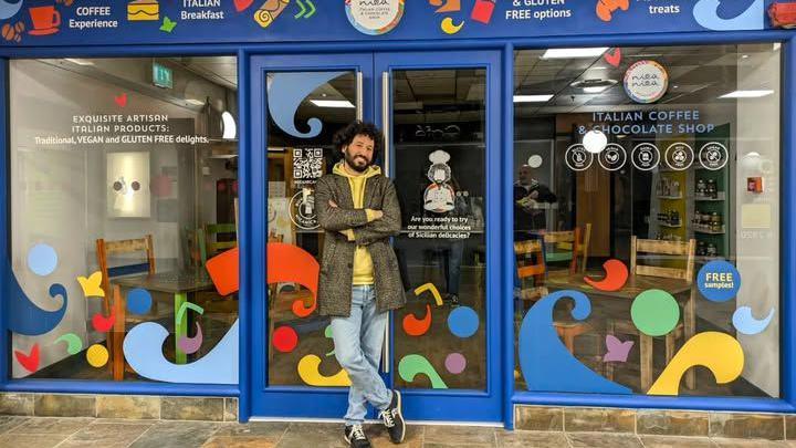 Giuseppe, a man who runs the Nica Nica café, standing in front of the shop front which has blue livery. Inside can be seen tables and chairs. 