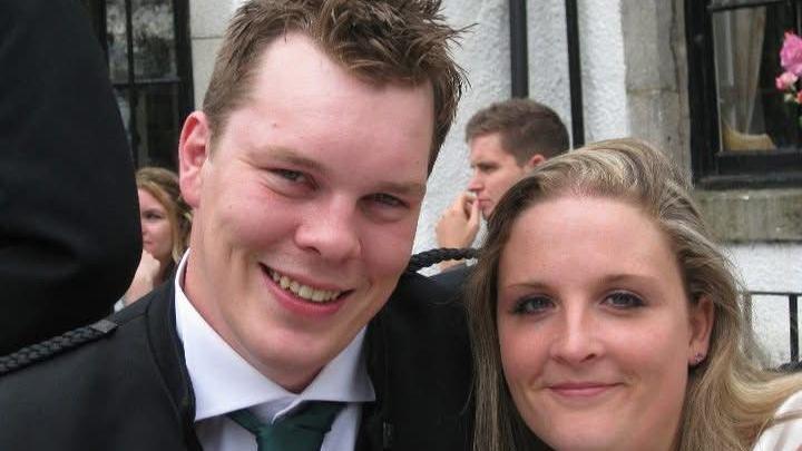 Felicity and James Hester are a couple in their thirties. They are both smartly dressed with James wearing a black jacket and tie. Only Felicity's head is visible but she has long blonde hair. He has his arm around her and they are both smiling.