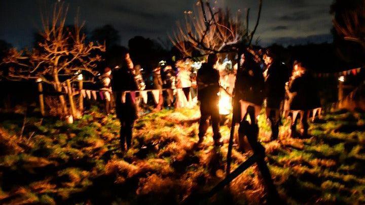 People stand around a fire in an apple orchard at night. Bunting hangs between the trees.