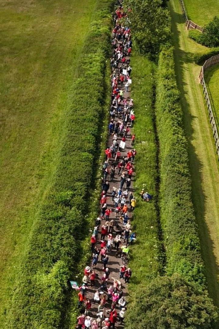 Hundreds of people are walking along a road in the middle of the countryside holding banners. The picture has been taken from a drone.