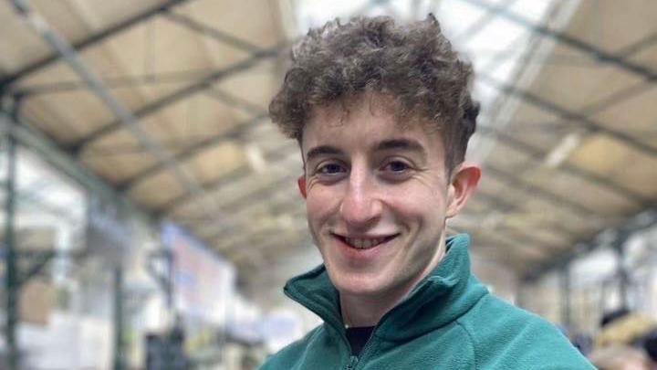 A man with short dark curly hair wearing a half-zip blue jumper.