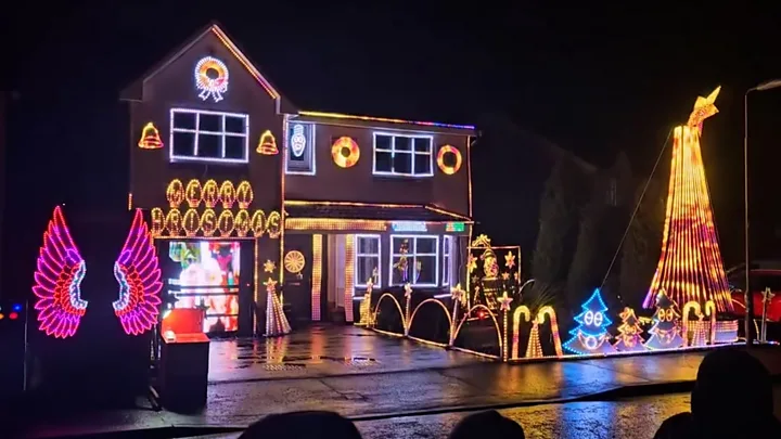 A two-storey house brightly illuminated in multi-coloured Christmas lights. There are small light up Christmas trees and light up candy canes around the border of the garden.