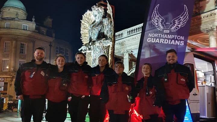 Seven volunteers from Northampton Guardians in their red uniforms standing outside All Saints Church with the Knife Angel, made up of thousands of knives, in the background.