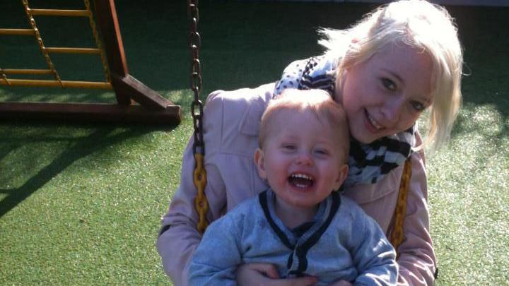 Bethany Vincent has white-blonde hair and is sitting on a swing on a sunny day, smiling at the camera. She's wearing a pink jacket and a black-and-white scarf. Her son Darren is sat on her lap and laughing.