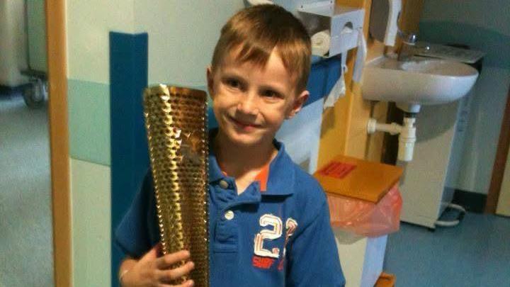 Finlay Thornell pictured as a child. He is in hospital. He has short brunette hair and is wearing a blue polo shirt and is holding the gold Olympic torch.He is looking at the camera and smiling