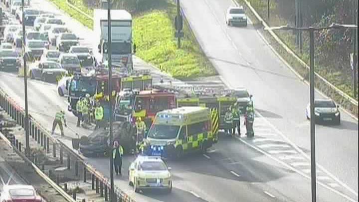 A motorway camera shows a number of emergency services vehicles. Traffic is backing up behind them. A car is on its roof. 