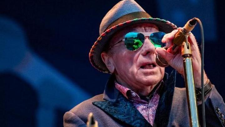 Van Morrison on stage. He is wearing a hat, a jacket and a patterned pink shirt. He is also wearing sunglasses and holding a microphone.