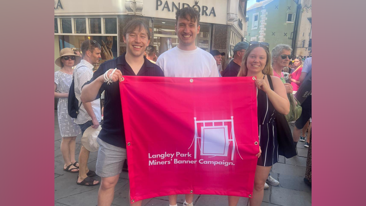 Three people stand on a busy street, smiling and holding a pink banner that reads: "Langley Park Miners' Banner Campaign". A blank miners' banner is also depicted on the print.