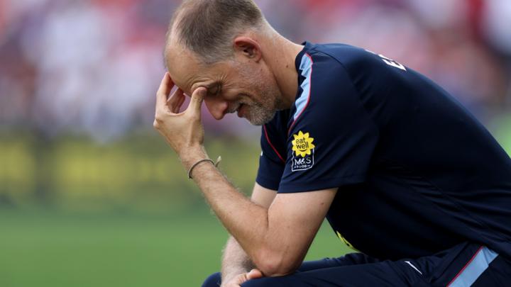 Thomas Tuchel has his left hand pressed to his forehead as he sits, face pointing downwards, wearing a blue England team training outfit